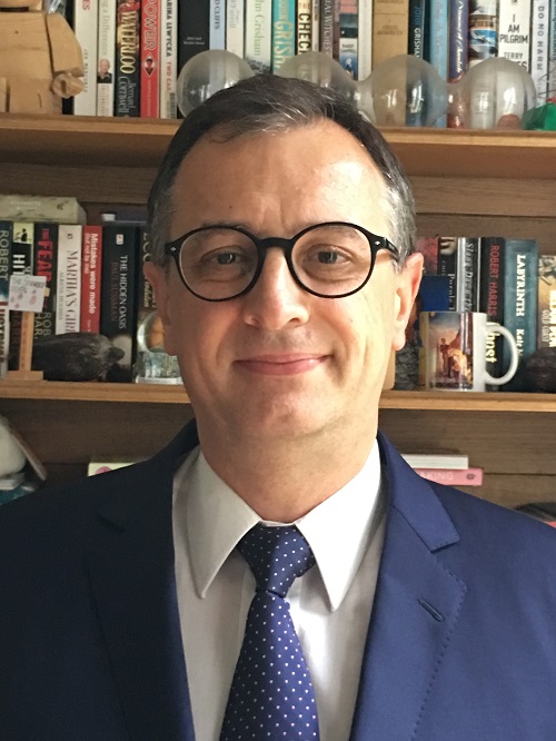 his book library; having a friendly smile on his face. He is wearing a navy blue suit jacket, white shirt, dotted blue tie.