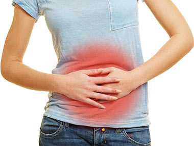 Woman wearing a light blue t-shirt and dark blue jeans with her hands pressed towards her abdomen. The area of the stomach is being highlighted in red in order to simulate pain.  Her face or bottom legs are not visible in the picture, the accent is on the abdomen.