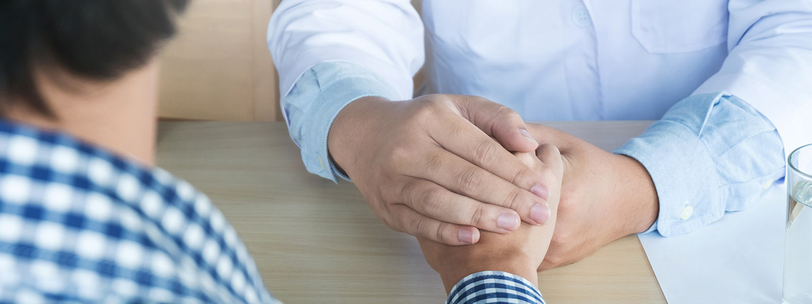 Surgeon and patient sited at the opposite sides of a desk; the surgeon holds patient’s hand, trying to reassure him about the success of the medical procedures